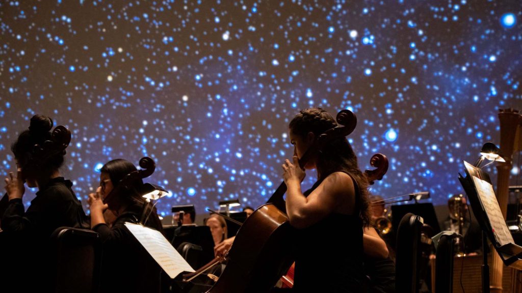 Orchestra members playing on stage. A starry sky shows on screen behind them. 